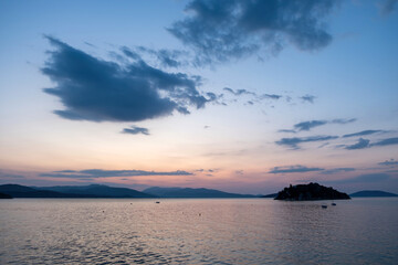 Sunset over Daskalio Island in Tolo Bay, Greece, calm sea water, silhouetted hills, and scattered clouds