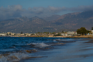 Waves reaching sandy beach with mountains and seaside town in the background in Crete, Greece