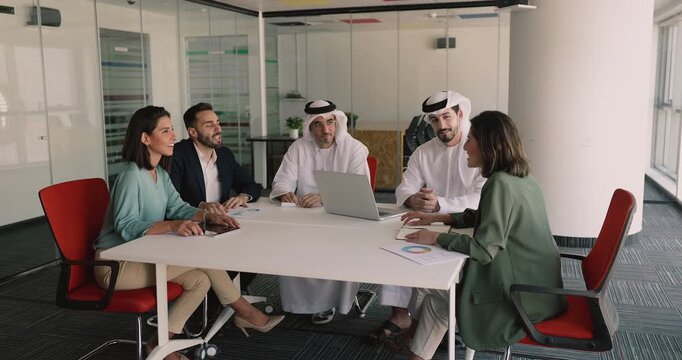 Multinational team engaged in discussion, exchanging ideas or solutions, analyze business metrics seated around conference table with laptop and printed papers displaying statistical data and charts - Powered by Adobe