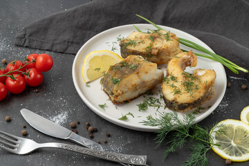 Baked zander fish with lemon, dill and cherry tomato on white ceramic plate with cutlery and linen serviette on brown stone board background.  Fried freshwater pike perch pieces.