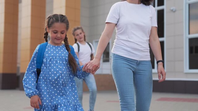 Mother walking holding daughter's hand while student girl with backpack smiles outside school building with parent nearby girl happy and confident walking together toward entrance near tiled facade