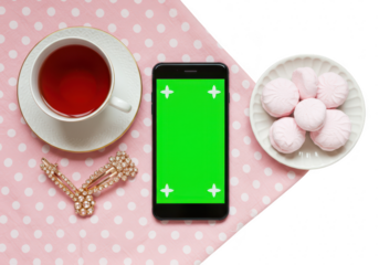 Top view of a cup of tea, a smartphone with a green screen, and marshmallows on a plate, placed on a pink polka dot fabric isolated on transparent background