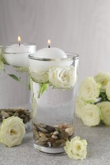 Burning candles in glasses with water, stones and rose flowers on light grey table, closeup