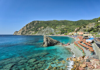 Obraz premium A view of the main beach at Monterosso al Mare, showing the vibrant turquoise water, a prominent rock formation, and a sandy shore filled with sunbathers and beach chairs