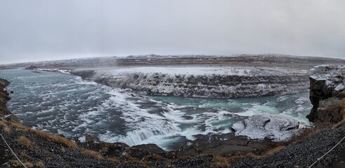 Panoramic view of Gullfoss waterfall in Iceland during winter, with powerful turquoise water flowing through snowy, rugged terrain under an overcast sky.