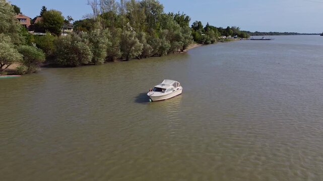A small motorboat is moored in a calm river, surrounded by trees in the distance. A drone makes a smooth half-circle flight around the boat