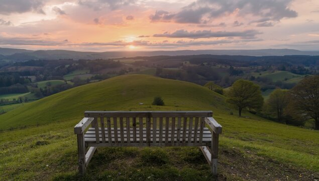 A wooden bench overlooks rolling green hills at sunset with a colorful sky in the background view
