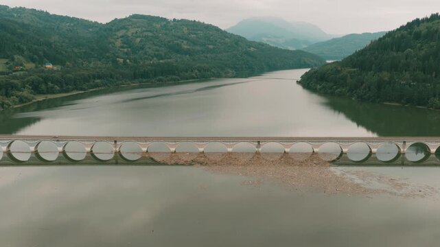 Aerial view of Bicaz Lake Bridge crossing calm waters with forested hills and mountains