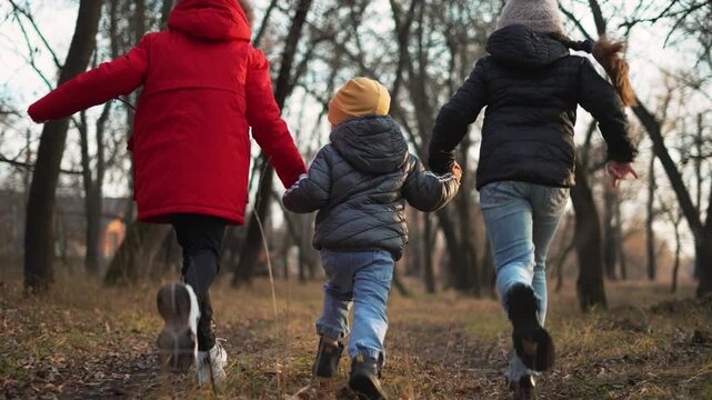 Three child run hand in hand through leafless park and forest in winter, wearing jacket and coat, sibling bond visible during outdoor play, family walk with joyful run and energetic movement