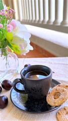 Black coffee cup with cookies and cherries sitting on a table with a small vase of flowers and a background of white columns enjoying morning sunlight,Black coffee, cookies, breakfast