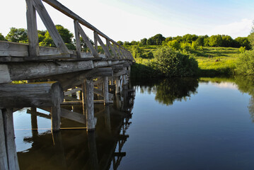 Scenic view of a wooden bridge crossing a river in summer. Countryside landscape with green trees, water, and blue sky. Rustic rural scene, travel, nature, and outdoor concept.
