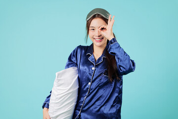 Smiling happy young Asian woman in pajama pillow showing ok gesture satisfied quality for night nap concept before she falls to sleep and enjoys a good sleep, headshots isolated studio blue background