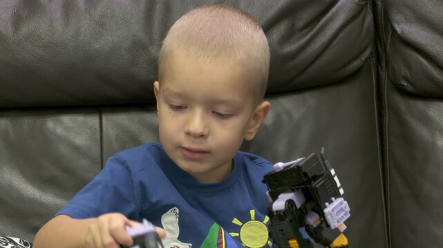 Child playing with new toy sitting on sofa at home. Concentrated face of little boy holding transformer robot toy. Three year old child examining new toy comments on his actions.