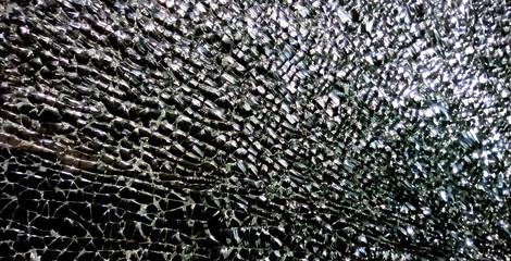 The photo captures a close-up of shattered glass, showcasing an intricate network of sharp fragments reflecting light. The dramatic texture and gleaming surfaces create a striking visual effect.
