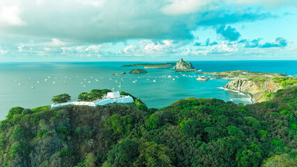 Fototapeta premium Vista aérea do Forte de Noronha e Baía de Santo Antônio ao pôr do sol, com barcos ancorados e vegetação densa compondo cenário tropical