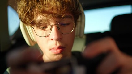 Close up of teenage boy with glasses wearing headphones and using smartphone in warm sunlight in the car - Powered by Adobe