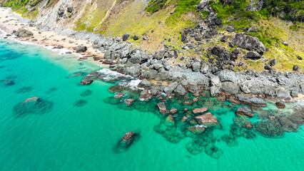 Visão aérea mostra ondas suaves se espalhando pela areia clara, enquanto rochas costeiras...