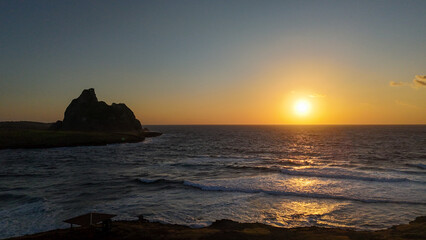 Fernando de Noronha ao amanhecer, com o nascer do sol refletindo no mar calmo e na paisagem litor&acirc;nea, revelando um espet&aacute;culo natural &uacute;nico e inspirador.