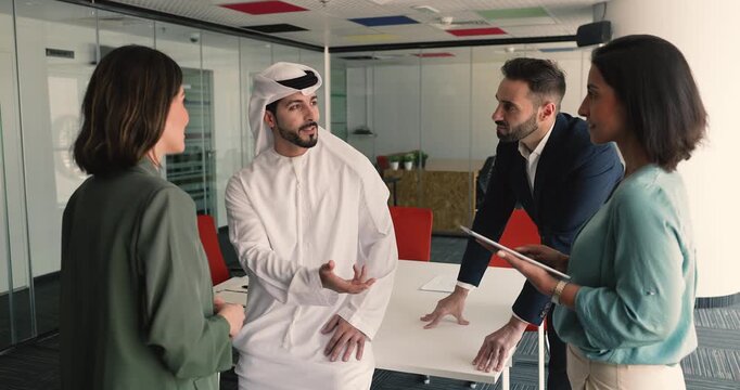 Young multicultural teammates engaged in briefing in meeting room. Arabic man dressed in traditional kandura lead professional discussion with Latin colleagues. Teamwork, ideas exchange, leadership