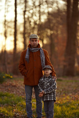 Obraz premium Man and boy wearing hats and jackets stand in a sunlit forest with hands in pockets. Concept of family, bonding, casual outdoor clothing. For family photo