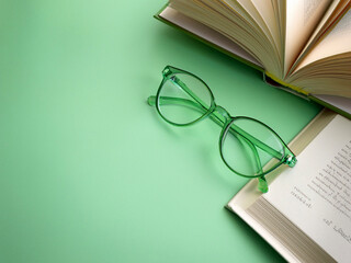Flat lay. Pair of women's reading glasses with a green frame and a book on a plain green background.
