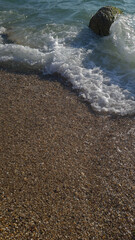 A close-up view of a sandy beach with gentle waves lapping at the shore. A large rock is partially submerged in the water.
