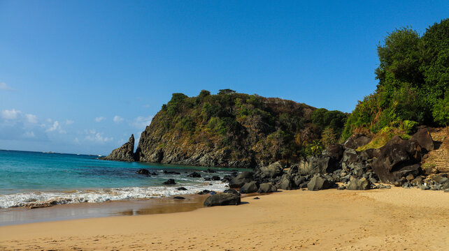 Praia em Fernando de Noronha com mar calmo, areia clara, morro em silhueta e luz suave do fim de tarde, mostrando cen&aacute;rio tropical, natureza preservada e turismo costeiro.