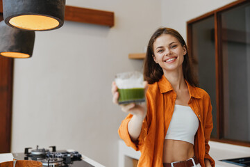 Young woman smiling and holding a green smoothie in a bright kitchen, wearing a stylish orange shirt, promoting healthy eating and a vibrant lifestyle