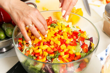 Hands preparing fresh vegetable salad in glass bowl. Chopped yellow and red bell peppers with cucumbers and lettuce. Healthy vegetarian food preparation. Natural ingredients for diet and nutrition.