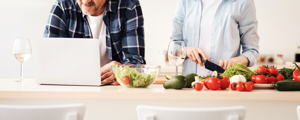 Couple enjoying and preparing healthy meal in kitchen and reading recipes on laptop. Happy adult woman makes salad of vegetables, man searches class in internet at kitchen interior, empty space