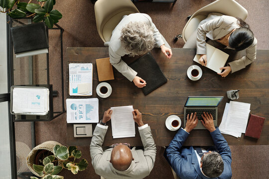 Top view showing group of middle aged multiethnic business professionals working together at conference table, analyzing documents, using laptop, writing notes, drinking coffee during meeting