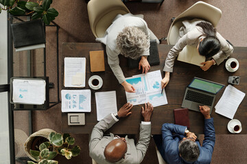 Diverse group of middle aged business professionals collaborating around office table, analyzing financial charts and graphs, using laptop and documents, top view workspace