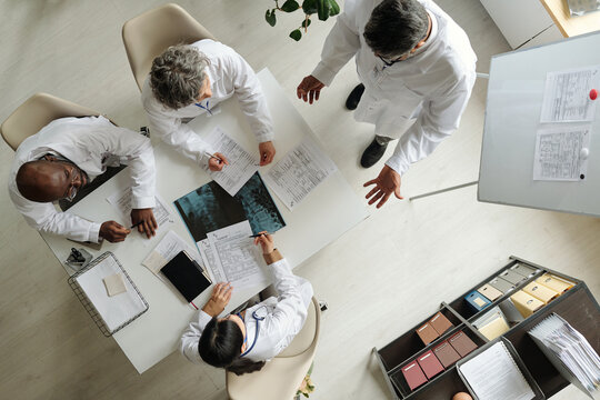 Group of middle aged multiethnic doctors discussing medical documents and analyzing x ray images around table in office, standing and sitting, collaborating on patient diagnosis - Powered by Adobe