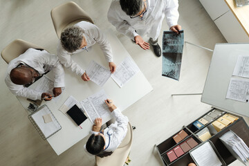 Group of middle aged and senior multiethnic doctors discussing medical records and examining X-ray image around table in hospital meeting room, top view showing collaborative teamwork