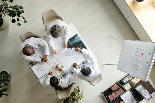Group of middle aged multiethnic doctors discussing medical documents around table, collaborating on patient care, reviewing charts and making notes, top view of healthcare professionals