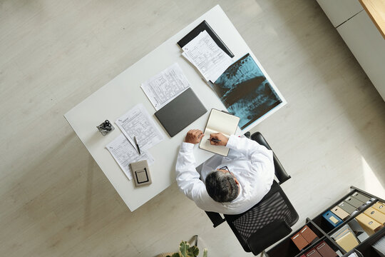 Middle aged Caucasian man sitting at desk reviewing medical documents and writing in notebook, analyzing X-ray image, top view showing organized workspace with paperwork and folders