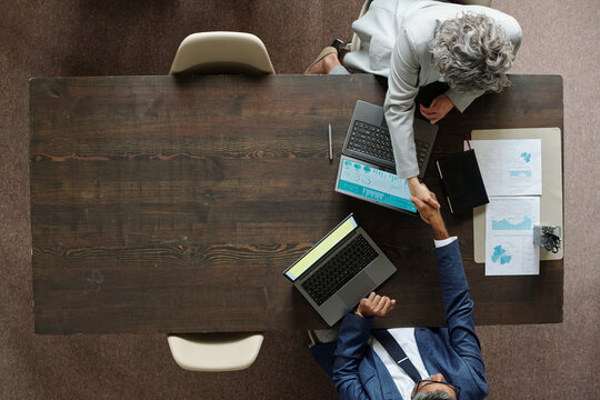 Top view showing middle aged Caucasian woman and middle aged Black man shaking hands across wooden table, both working on laptops with business charts and documents visible