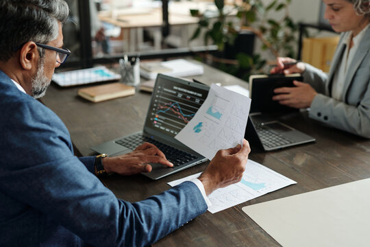 Middle aged Caucasian man analyzing financial charts on laptop while holding printed graphs, sitting at desk with middle aged Caucasian woman using digital tablet in office workspace