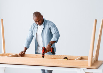 Professional african american craftsman drilling wooden planks of table, assembling new furniture and screwing details of desk with electric screwdriver, free space