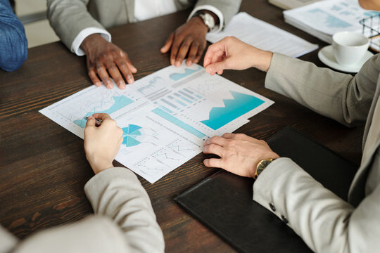 Caucasian woman and Black man discussing financial charts and graphs during business meeting, hands gesturing over printed documents on wooden table, top view composition - Powered by Adobe