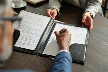 Middle aged Caucasian man signing contract while middle aged Caucasian woman explaining document details, both sitting at wooden table in office, top view showing hands and paperwork