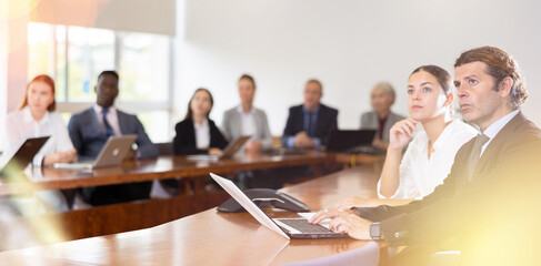 Absorbed mature white dark-haired business man in formal suit sitting with his young white female colleague apart in conference room and intently listening to speaker's presentation during corporate