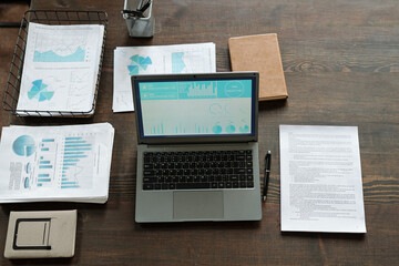 Obraz premium Top view showing open laptop displaying charts and graphs on screen, surrounded by printed financial reports, pen, notebook, and office supplies on wooden desk