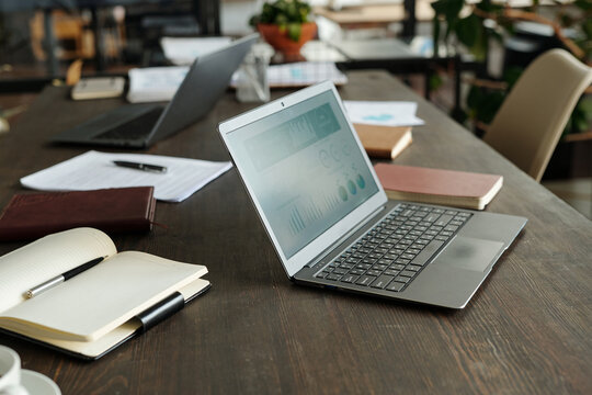 Top view showing open laptop displaying business analytics on wooden desk surrounded by notebooks, documents, pen and another laptop, workspace arranged for office work or remote meeting