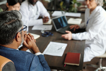 Middle aged Caucasian man sitting at table with diverse group of doctors discussing medical documents and laptop during meeting, hands clasped under chin, top view