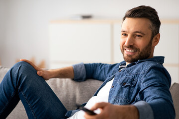 Fototapeta premium Watching TV at home. Happy middle-aged man having rest, switching channels with remote controller and smiling, empty space, closeup. Bearded man enjoying his weekend, watching TV in living room