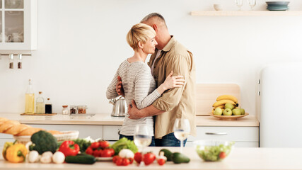 Fun, celebrate anniversary, date and enjoy romance together. Bright fresh vegetables for salad, glasses of light wine on table. Happy mature man and woman dancing on kitchen interior, empty space
