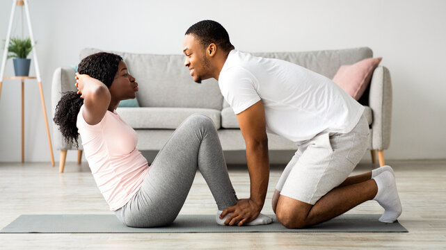 Athletic black couple exercising together at home, working on abs. Cheerful african american man in sportswear husband holding his girlfriend or wife legs while she is doing exercises for belly
