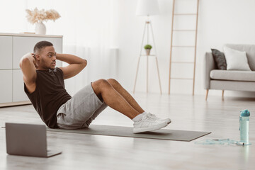 Side View Of African American Guy Doing Sit-Ups Exercising At Laptop Sitting On Floor At Home. Man...