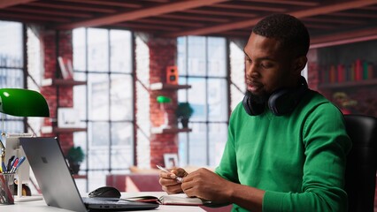 African american person at home extracting information on textbook, writing down data on notepad and identifying trends. Guy compiling analytical reports and taking notes. Camera A.